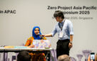 Tanzila Khan holding her board game and speaking into the mic held by a volunteer on stage.