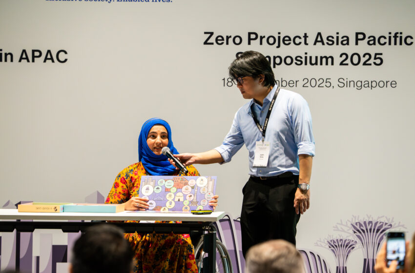 Tanzila Khan holding her board game and speaking into the mic held by a volunteer on stage.
